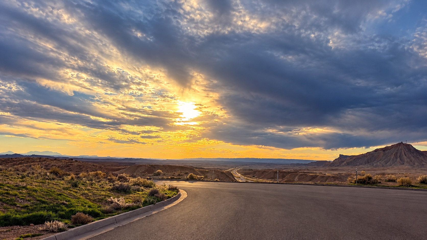 Sunlight breaking through blue-gray clouds above a desert highway and rugged mesa.