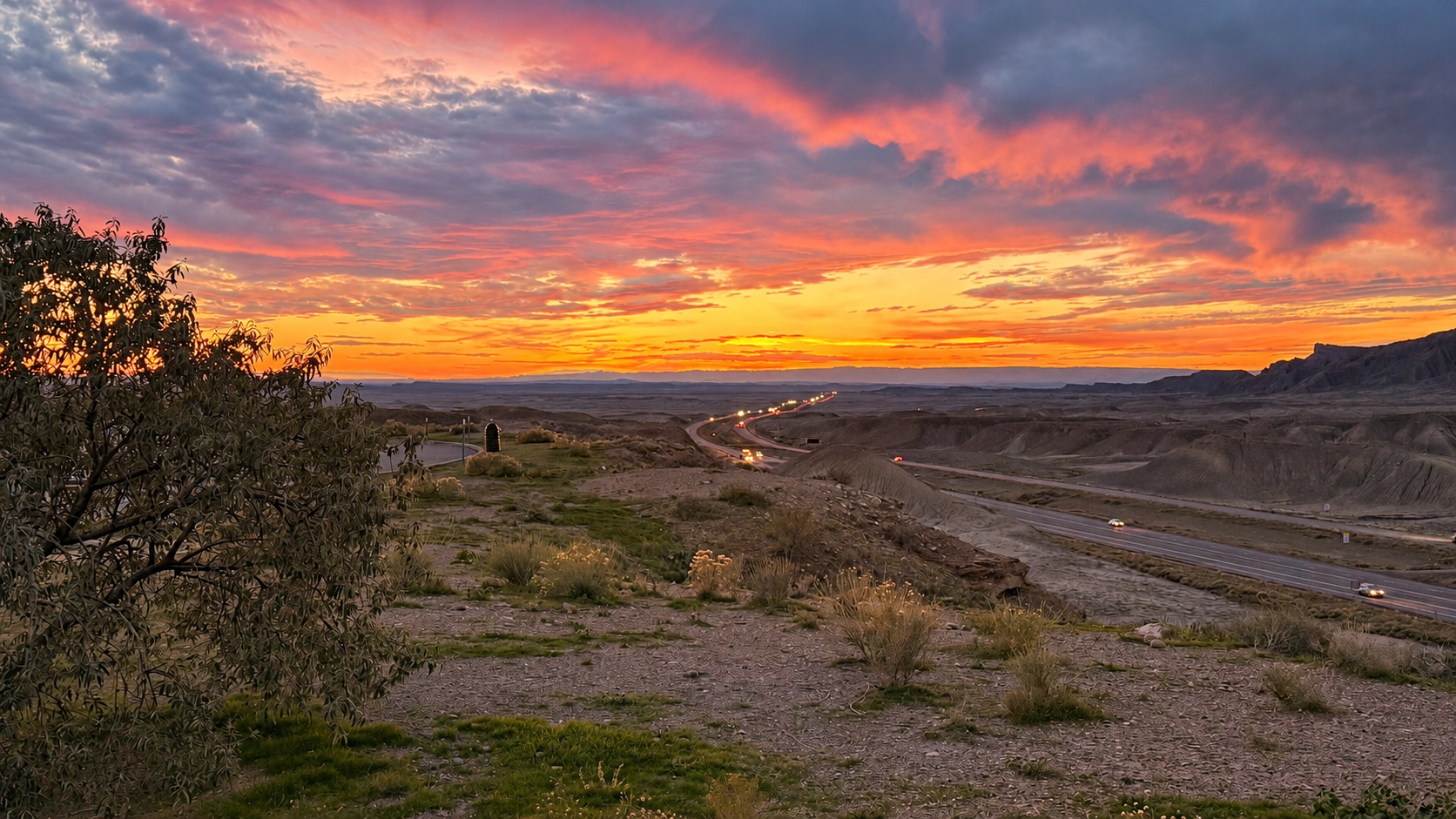 Pink and orange sunset clouds above a winding highway through desert hills, framed by a tree.