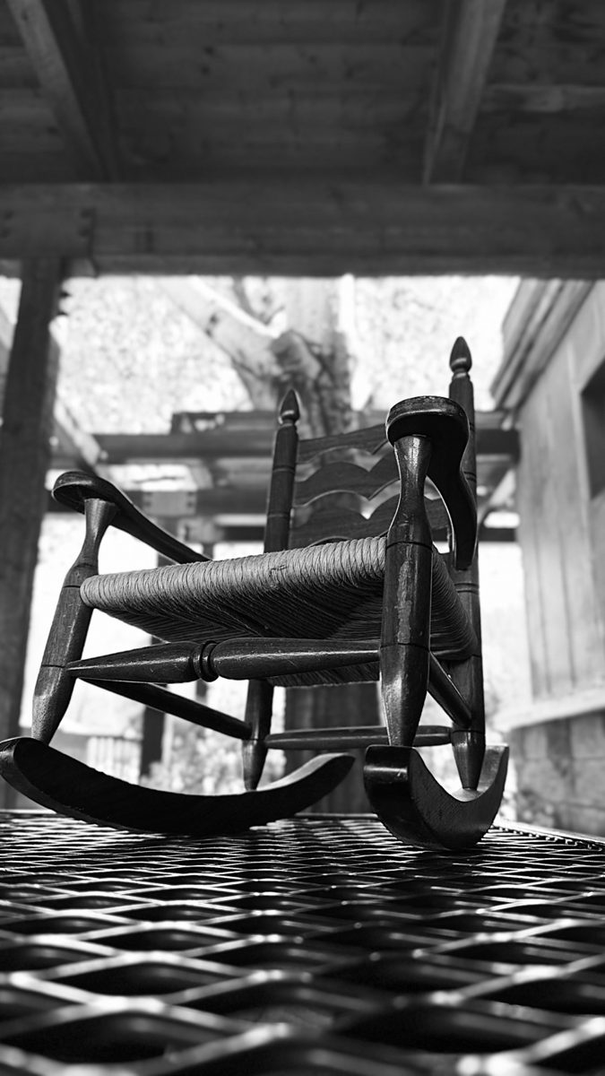 Black-and-white photograph of a rocking chair beneath a shaded porch.