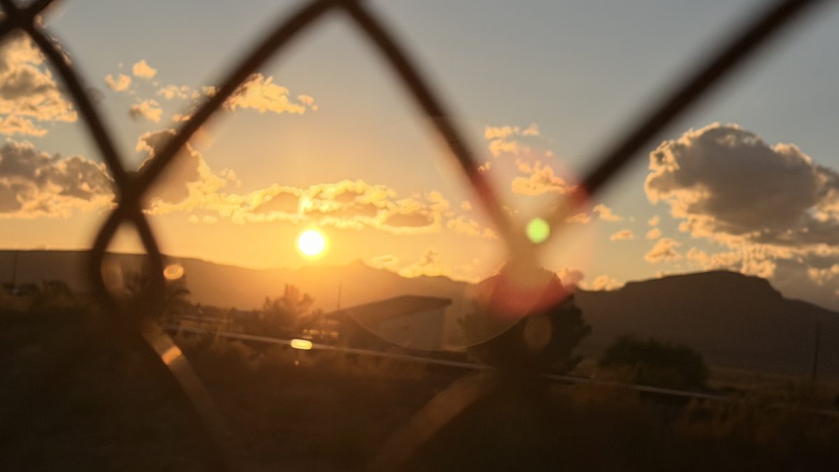 Sunset over distant mountains seen through a chain-link fence.